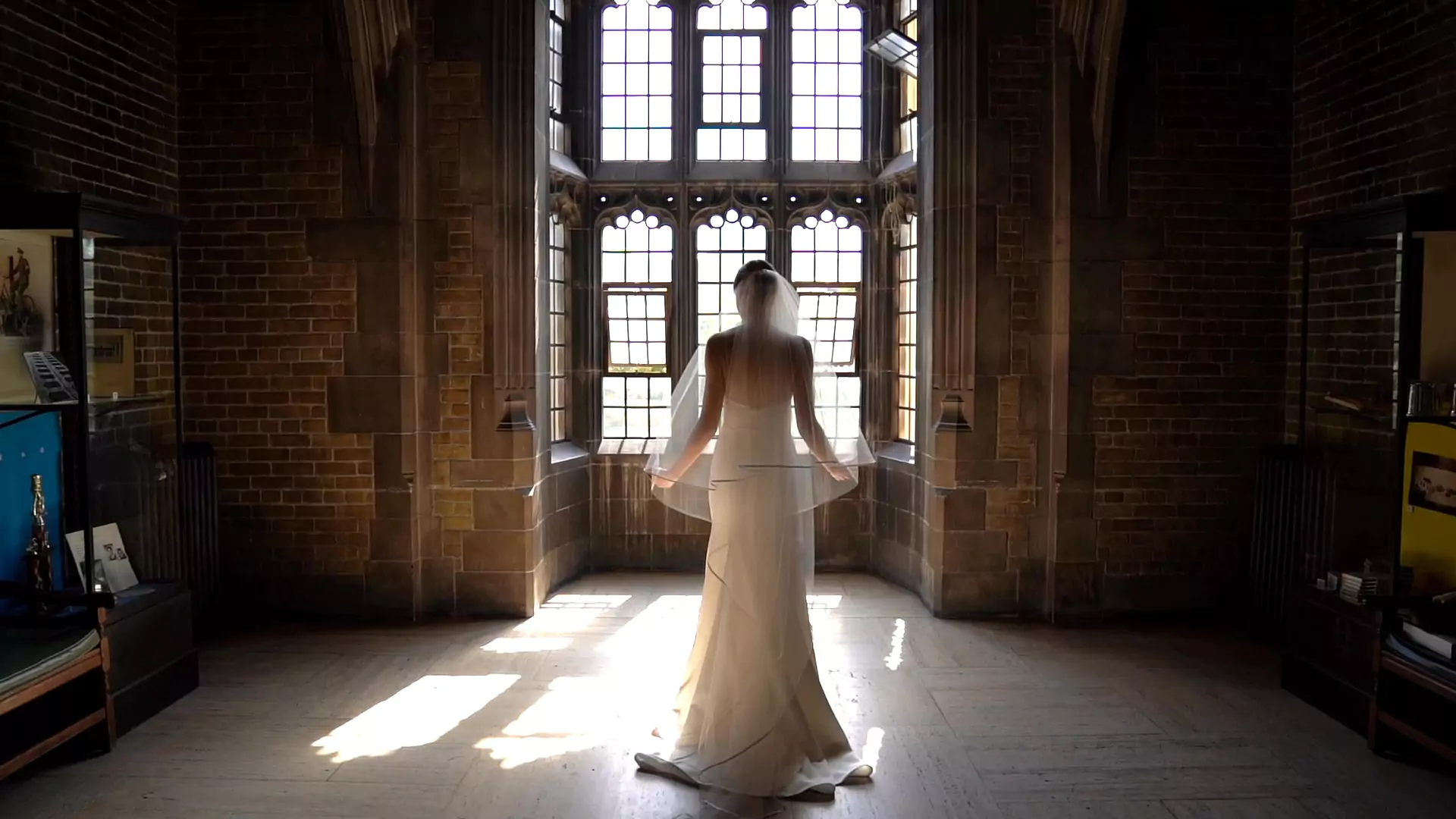 Jennika facing away from the camera infront of a window inside Hart House, beautifully backlit with sun shining through her long veil.